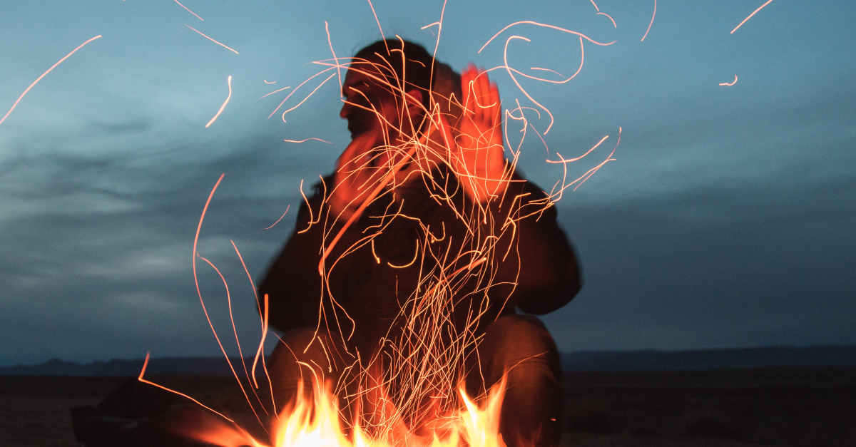 man sitting so close to flames in fire outdoors at night sparks flying hands up